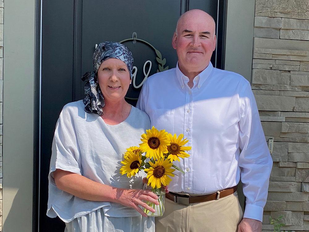 Pam wearing a head scarf and holding flowers, standing next to her husband